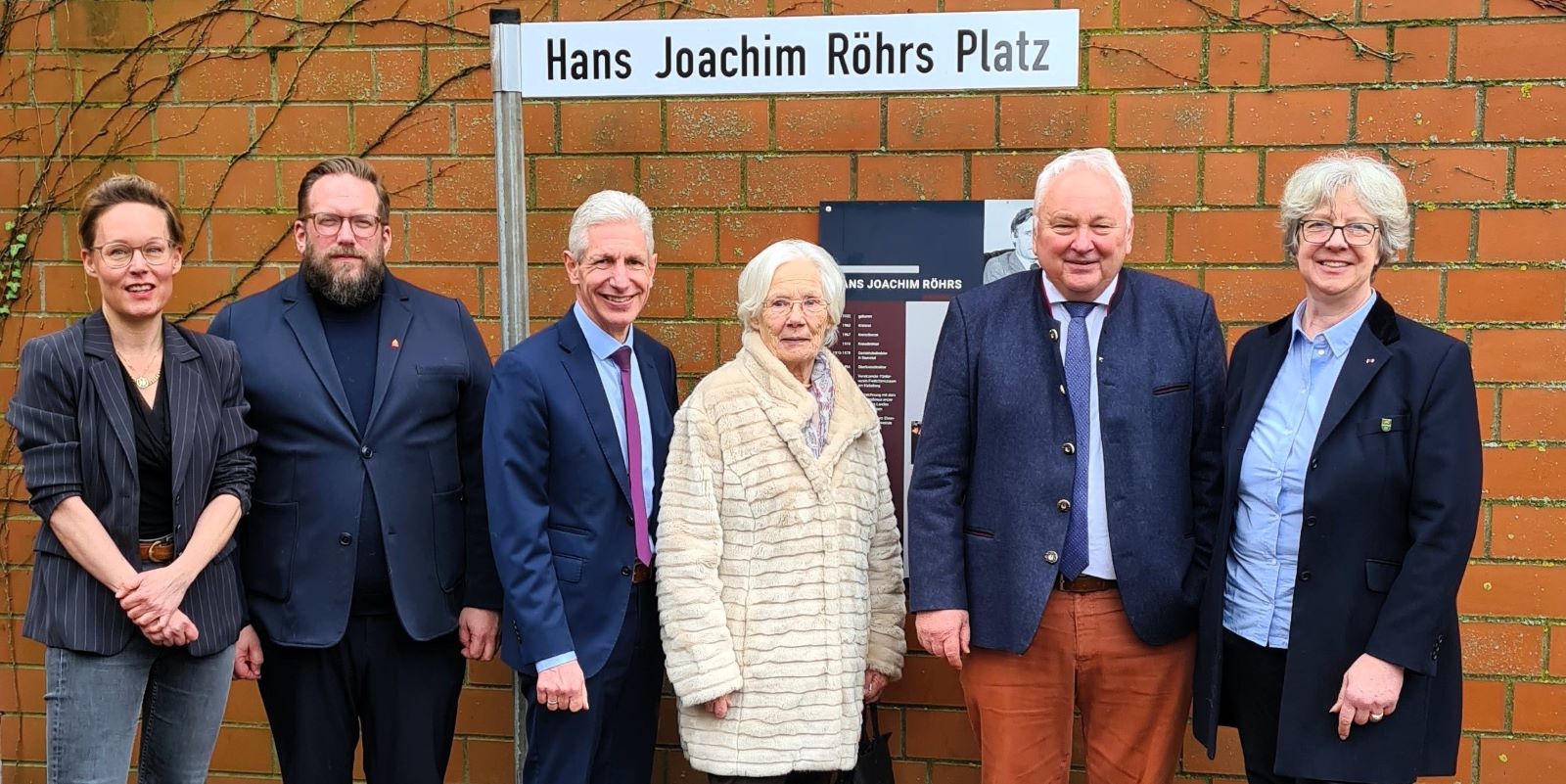 Vor dem Schild des eingeweihten „Hans Joachim Röhrs Platzes“ im Freilichtmuseum am Kiekeberg: Carina Meyer (v.l.) und Stefan Zimmermann (Kaufmännische Geschäftsführerin und Direktor), Rainer Rempe (Landrat), Anneke Korf (Schwester von H. J. Röhrs), Heiner Schönecke und Emily Weede (Bürgermeisterin Gemeinde Seevetal). Foto: FLMK